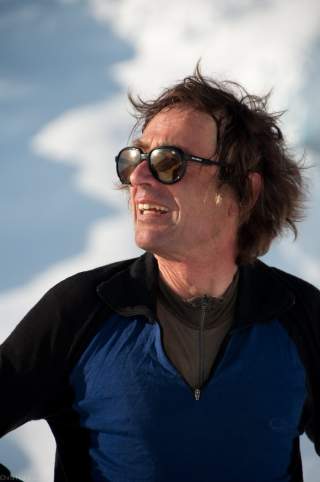 Mann mit zerzaustem Haar trägt eine dunkle Sonnenbrille und ein blau-schwarzes Oberteil, im Hintergrund verschwommene Schneelandschaft.