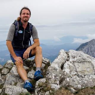 Mann mit Rucksack und Wanderkleidung sitzt auf Felsen in bergiger Landschaft bei bewölktem Himmel.
