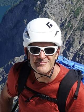 Mann mit weißem Kletterhelm, Sonnenbrille und rotem T-Shirt vor einer felsigen Bergkulisse.