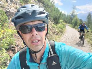 Zwei Männer tragen Helme und fahren Mountainbikes auf einem schmalen, bergigen Waldweg bei sonnigem Wetter. Einer macht eine Nahaufnahme, während der andere im Hintergrund fährt.