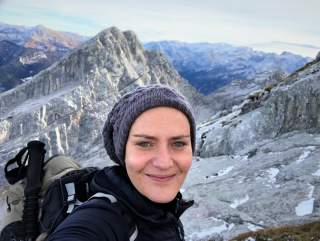 Frau mit Mütze und Wanderrucksack macht Selfie vor schneebedeckten, felsigen Berggipfeln bei klarem Himmel.