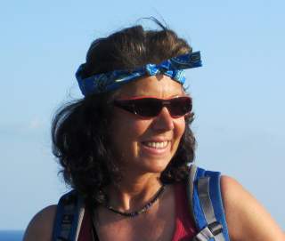 Frau mit braunen lockigen Haaren, blauer Bandana, Sonnenbrille und blauer Tagesrucksack vor blauem Himmel, die lächelt.