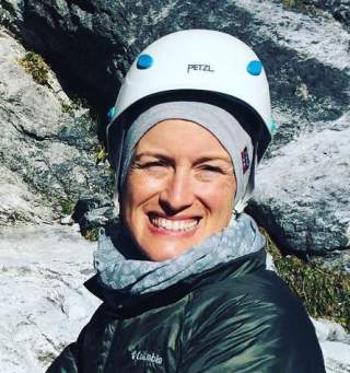 Porträt einer lächelnden Frau mit Kletterhelm und Outdoor-Bekleidung vor felsigem Hintergrund.