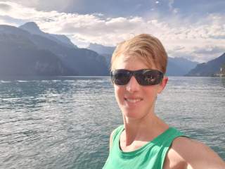 Frau mit kurzen blonden Haaren und Sonnenbrille posiert in grünem Top vor einem See mit Bergen im Hintergrund an einem sonnigen Tag.