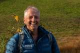 Ein lächelnder Mann mit grauen Haaren trägt eine blaue Jacke und einen Rucksack vor einer grünen Wiese.