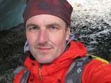 Porträt eines Mannes mit roter Jacke, rotem Stirnband und grauem Rucksack in einer felsigen Outdoor-Umgebung.