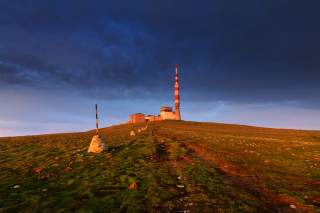 Ein Gebäudekomplex mit einem rot-weiß gestreiften Sendemast steht auf einem grasbewachsenen Hügel unter einem dunklen, wolkigen Himmel bei Sonnenuntergang. Ein Weg führt vom Vordergrund direkt zu dem Gebäude.