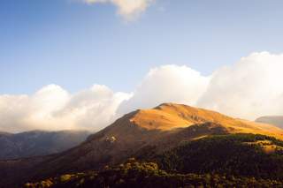 Berglandschaft mit sonnendurchfluteten Hügeln und Nadelwäldern unter blauem Himmel mit weißen Wolken.