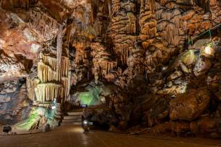 Innenansicht einer beleuchteten Tropfsteinhöhle mit Stalagmiten, Stalaktiten und einem gepflasterten Weg.