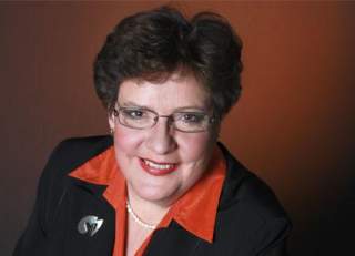 Porträt einer Frau mit kurzen braunen Haaren, die eine Brille trägt und in einem schwarzen Blazer mit orangem Kragen vor einem braunen Hintergrund lächelt.