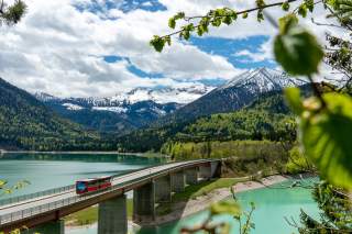 Ein roter Bus fährt über eine Brücke, die einen türkisfarbenen See verbindet, umgeben von grünen Wäldern und schneebedeckten Bergen unter einem bewölkten Himmel.