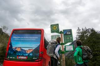 Zwei Menschen stehen an Bushaltestelle mit rotem Bus