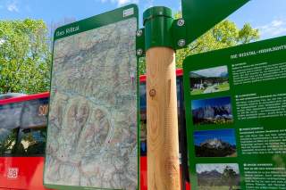 Informations- und Wanderkarte des Risstals mit Highlights, daneben ein roter Bus und grüne Bäume im Hintergrund.