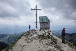 Gipfelkreuz und kleine Holzhütte auf einem Berggipfel mit zwei wandernden Personen und bewölktem Himmel im Hintergrund.