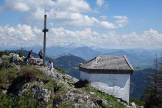 Drei Personen sitzen auf einem felsigen Berggipfel neben einem Holzmast und einem kleinen weißen Gebäude mit Holzdach, im Hintergrund sind bewaldete Berge und schneebedeckte Gipfel zu sehen.