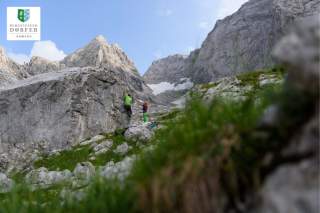 Zwei Personen klettern an einem großen Felsen in einer bergigen Landschaft mit schroffen Gipfeln und etwas Schneeresten, unter blauem Himmel.