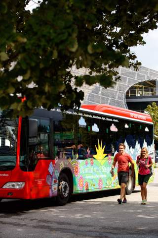 Ein bunter DB Oberbayernbus steht auf einer Straße, daneben gehen ein Mann und eine Frau in Wanderkleidung, im Vordergrund sind Blätter eines Baumes sichtbar.