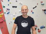 Ein lächelnder Mann steht vor einer Kletterwand mit bunten Griffen in einer Kletterhalle. Er trägt ein schwarzes T-Shirt mit einer Berggrafik.