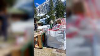 Große weiße Säcke mit rotem Band, gefüllt mit verschiedenen Materialien, stehen im Freien vor schneebedeckten Bergen und Nadelbäumen, daneben eine Person in schwarzer Kleidung.