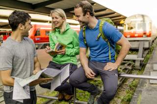 Drei Wanderer mit Outdoor-Bekleidung und Rucksäcken unterhalten sich fröhlich am Bahnsteig, während ein Zug im Hintergrund hält. Zwei von ihnen sitzen auf einem Geländer, einer steht mit einer Karte in der Hand.