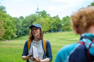 Frau mit Brille, Kappe und Rucksack steht auf einer Wiese im Park, hält ein Notizbuch und spricht; im Vordergrund unscharf eine weitere Person, im Hintergrund Bäume und ein Turm.