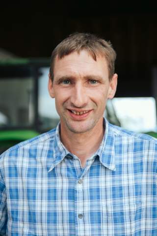 Das Bild zeigt den Bergbauern Konrad Wörndl. Er steht vor seinem grünen Traktor und trägt ein blau-weiß kariertes Hemd.