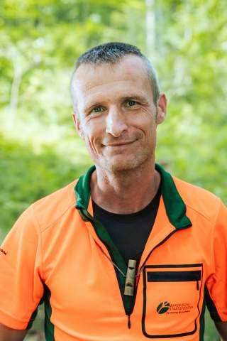 Ein Mann trägt seine orange-grünen Dienstkleidung mit dem Logo der Bayerischen Staatsforsten auf der Brusttasche. Er steht im Wald, umgeben von Bäumen. Er blickt lächelnd in die Kamera.