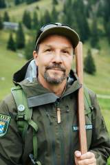Der Ranger Alex Römer steht in seiner grünen Dienstkleidung auf einer Bergwiese. In der Hand hält er einen Holzstab.
