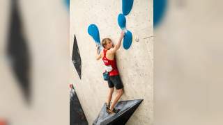 Junger Kletterer in rotem Trikot und Kletter-Equipment erklimmt eine Indoor-Kletterwand mit blauen Griffen.