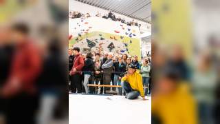 Menschenmenge steht und sitzt vor einer Boulderwand, während ein Mann in gelber Kapuzenjacke mit Mikrofon hockt und spricht.