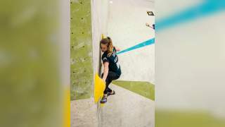 Eine junge Frau klettert an einer Kletterwand und hält sich dabei an großen gelben Griffen fest. Sie trägt Kletterschuhe und ein schwarzes Shirt mit der Aufschrift „TEAM BAY“.