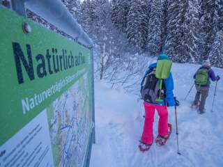 Zwei Personen mit Schneeschuhen wandern auf einem verschneiten Waldweg, im Vordergrund steht ein grünes Schild mit Informationen zur Naturverträglichkeit der Skitour.