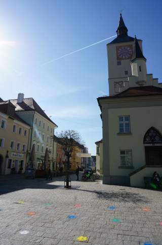 Stadtplatz mit alten Gebäuden und einem Uhrturm bei klarem Himmel, bunte Blumenmuster auf dem gepflasterten Boden.