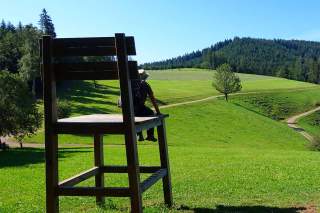 Eine Person mit Sonnenhut sitzt auf einem überdimensional großen Holzstuhl auf einer grünen Wiese mit Hügeln und Bäumen im Hintergrund bei blauem Himmel.