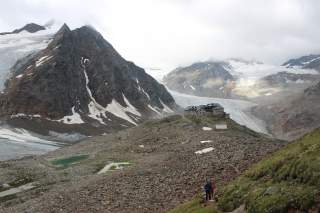 Berglandschaft mit teils schneebedeckten Gipfeln und Gletscher, in der Bildmitte eine Berghütte auf einem Felsvorsprung, im Vordergrund zwei Wanderer auf einem Pfad.