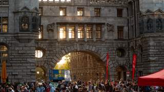 Menschenmenge vor einem historischen Gebäude mit großem Steinbogen, durch den Sonnenlicht scheint, bei einer Veranstaltung mit roten Fahnen und einem roten Zelt.