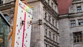 Zwei Personen klettern an einer mobilen Kletterwand vor einem historischen Gebäude. Die Kletterwand ist mit roten Griffen und LED-Leuchten ausgestattet.