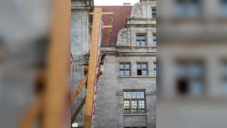 Zwei Personen klettern an einer Outdoor-Kletterwand mit Sicherheitsseilen vor einem alten steinernen Gebäude mit vielen Fenstern.