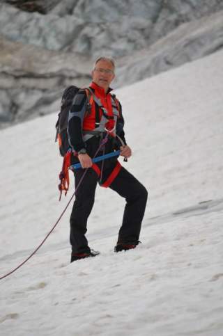Ein Bergsteiger in roter und schwarzer Ausrüstung steht mit Steigeisen und Eispickel auf einem schneebedeckten Hang, verbunden mit einem Seil.