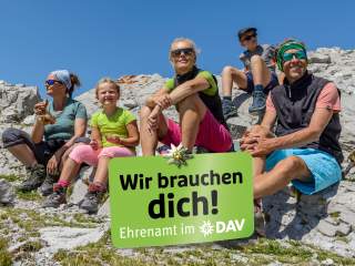 Fünf Personen unterschiedlichen Alters sitzen in Wanderkleidung auf felsigem Gelände unter blauem Himmel, davor ein grünes Schild mit der Aufschrift „Wir brauchen dich! Ehrenamt im DAV“.