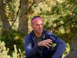 Mann mit rosafarbenem Stirnband und Sonnenbrille sitzt entspannt im Wald und gestikuliert. Er trägt eine blaue Jacke und spricht scheinbar.