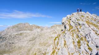 Zwei Wanderer stehen auf einem felsigen Berggipfel mit Blick auf eine weite, zerklüftete Berglandschaft unter blauem Himmel.