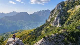 Ein Mensch steht auf einem Felsen mit Blick auf bewaldete Berge und einen klaren Himmel. Im Hintergrund sind klein weitere Menschen auf einem Pfad zu sehen.