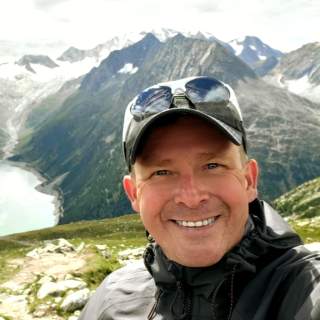 Porträt eines lächelnden Mannes mit Baseballcap und Sonnenbrille vor einer Berglandschaft mit schneebedeckten Gipfeln und einem See.
