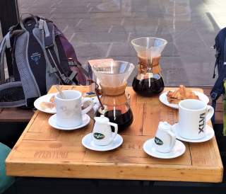 Ein Holztisch mit zwei Kaffeekaraffen, zwei Tassen, je einer Zuckerdose, je einem Teller mit Croissants und zwei Rucksäcken im Hintergrund.