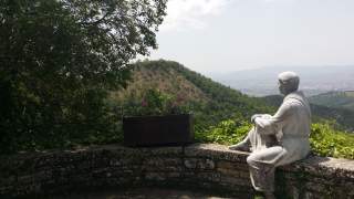 Statue einer sitzenden Person blickt auf bewaldete Hügel und eine offene Landschaft im Hintergrund.