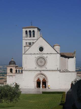 Vorderansicht einer alten, weißen Steinbasilika mit einem großen, kunstvoll verzierten Rundfenster und einem Glockenturm unter blauem Himmel. Vor der Kirche liegt ein gepflegter Rasen mit Hecken.