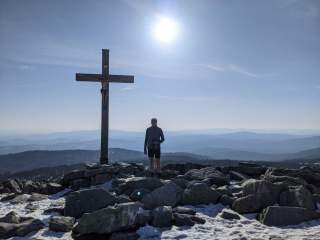 Person steht bei Sonnenschein auf einem schneebedeckten Berggipfel neben einem großen Holzkreuz mit einer Christusfigur, im Hintergrund sind entfernte Bergketten sichtbar.