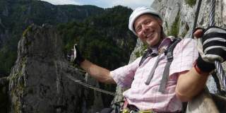 Ein lächelnder Mann mit Helm und Kletterausrüstung hält sich an einem Stahlseil an einer Felswand fest, im Hintergrund sind Berge und Wald zu sehen.