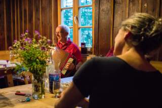 Ein Mann spielt in einem rustikalen Raum Akkordeon, während eine Frau ihm aufmerksam zuhört. Auf dem Tisch im Vordergrund stehen Blumen, Getränke und verschiedene Gegenstände.